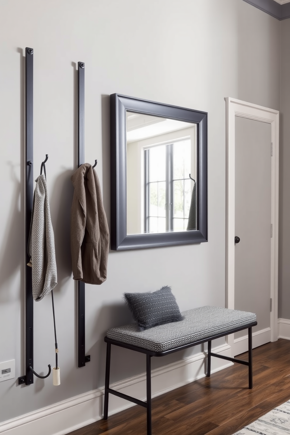 A stylish winter hallway features wall-mounted coat racks made of sleek metal with a matte black finish. The walls are painted in a soft gray hue, creating a warm and inviting atmosphere. Beneath the coat racks, a cozy bench upholstered in a textured fabric offers a perfect spot to sit and remove shoes. A large, framed mirror hangs above the bench, reflecting natural light and enhancing the space's openness.