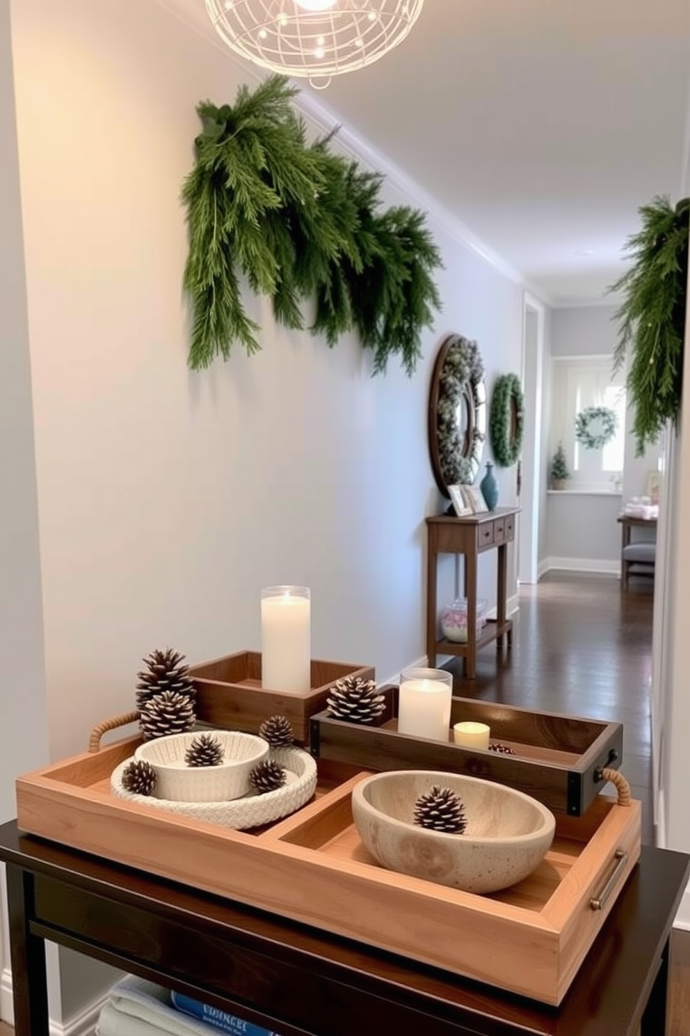 A collection of decorative trays in various sizes is arranged on a console table in the hallway. The trays are made of natural wood and ceramic, showcasing a mix of textures while holding seasonal decor items like pinecones and small candles. The hallway is adorned with winter-themed decorations, including a garland of evergreen branches draped along the wall. Soft white lights twinkle above, creating a warm and inviting atmosphere for guests as they enter the home.