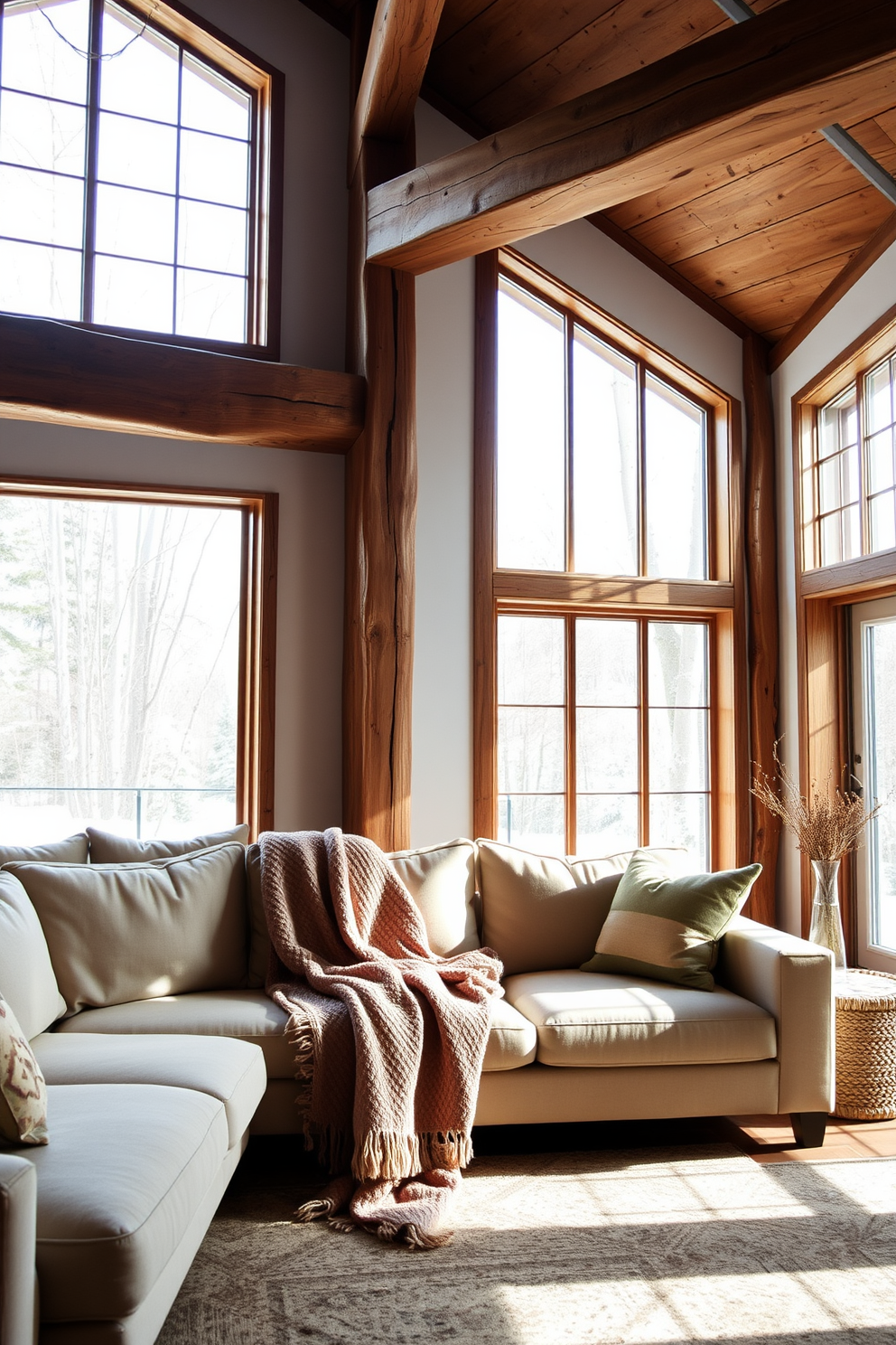 A cozy winter loft living room features a plush sofa adorned with a warm throw blanket draped casually over one arm. Large windows allow natural light to flood the space, highlighting the rustic wooden beams and soft, neutral color palette.