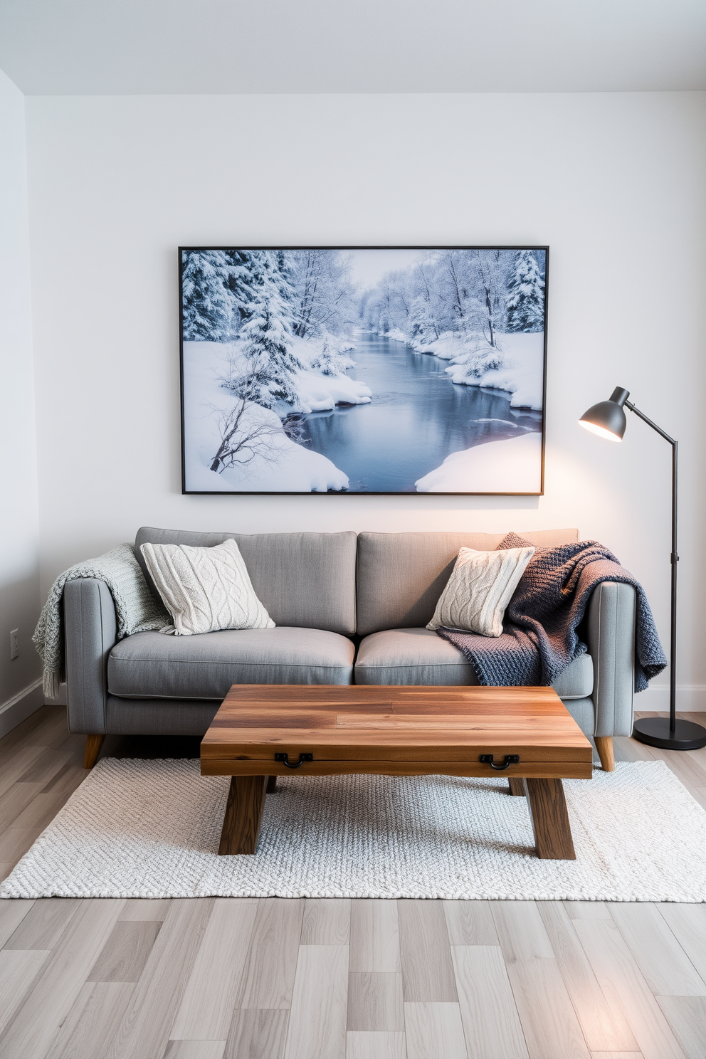 A serene winter landscape artwork hangs on the wall, showcasing snow-covered trees and a tranquil river. The color palette consists of soft blues and whites, creating a calming atmosphere. In the small living room, a cozy arrangement features a plush gray sofa adorned with knitted throw blankets. A rustic wooden coffee table sits at the center, surrounded by warm lighting from a stylish floor lamp.